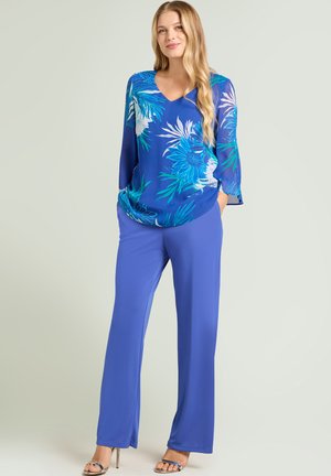 Kobieta z długimi blond włosami, ubrana w niebieską bluzkę w kwiaty, dopasowane niebieskie spodnie oraz srebrne sandały na obcasie z odkrytymi palcami, stojąca z rękami w kieszeniach.