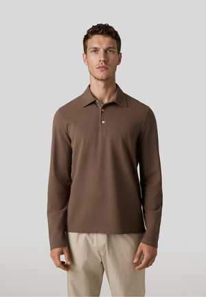 Man draagt een bruin poloshirt met lange mouwen en beige broek, staand tegen een effen lichtgrijze achtergrond.