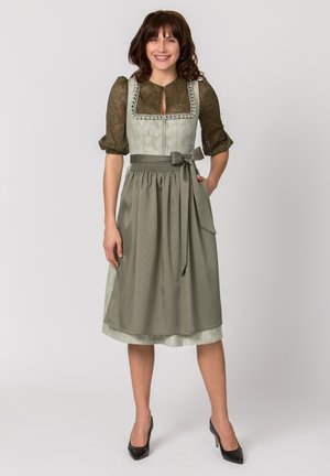 Woman smiling, standing, wearing olive green dirndl dress with lace blouse and tied apron, paired with black high heels, plain background.