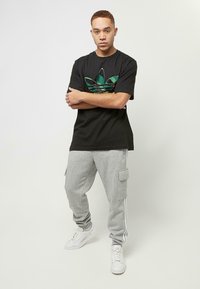 Zwarte katoenen T-shirt met groen Adidas-logo, gecombineerd met grijze joggingbroek met zijzakken en witte streepelementen, en witte sneakers.