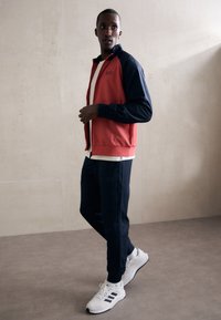 Veste à zip rouge et marine, t-shirt blanc en dessous, pantalon de jogging marine, et baskets blanches avec des accents noirs. Tissu lisse, coupe athlétique.