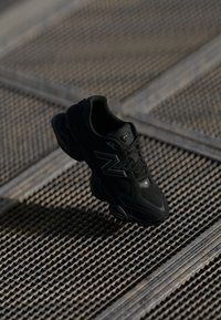 Baskets de sport noires avec une tige en maille et en matière synthétique, semelle rembourrée et accents réfléchissants, placées au-dessus d'une surface texturée.