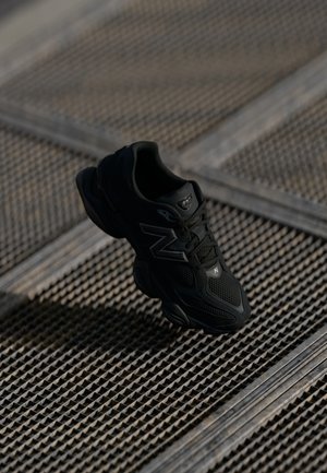 Black athletic shoe with mesh and synthetic upper, cushioned sole, and reflective accents, positioned above a textured surface.