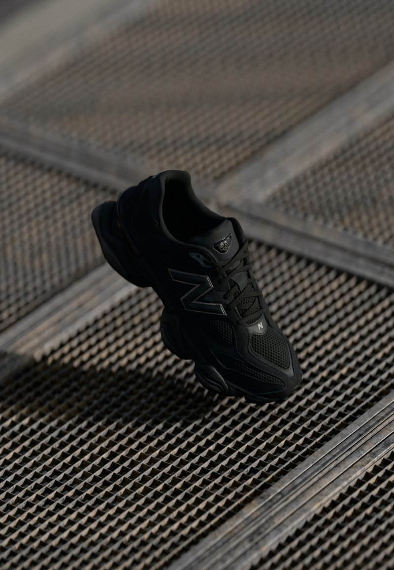 Baskets de sport noires avec une tige en maille et en matière synthétique, semelle rembourrée et accents réfléchissants, placées au-dessus d'une surface texturée.