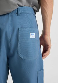 Blauwe werkbroek met een gestructureerde afwerking, voorzien van een patchzak met een wit label. De tailleband is aansluitend en iets zichtbaar.