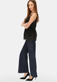 Czarna bluzka z koronkową lamówką i falowanym dołem, zestawiona z wysokim stanem, szerokimi nogawkami ciemnych jeansów oraz czarnymi szpilkami o wydłużonych czubkach.