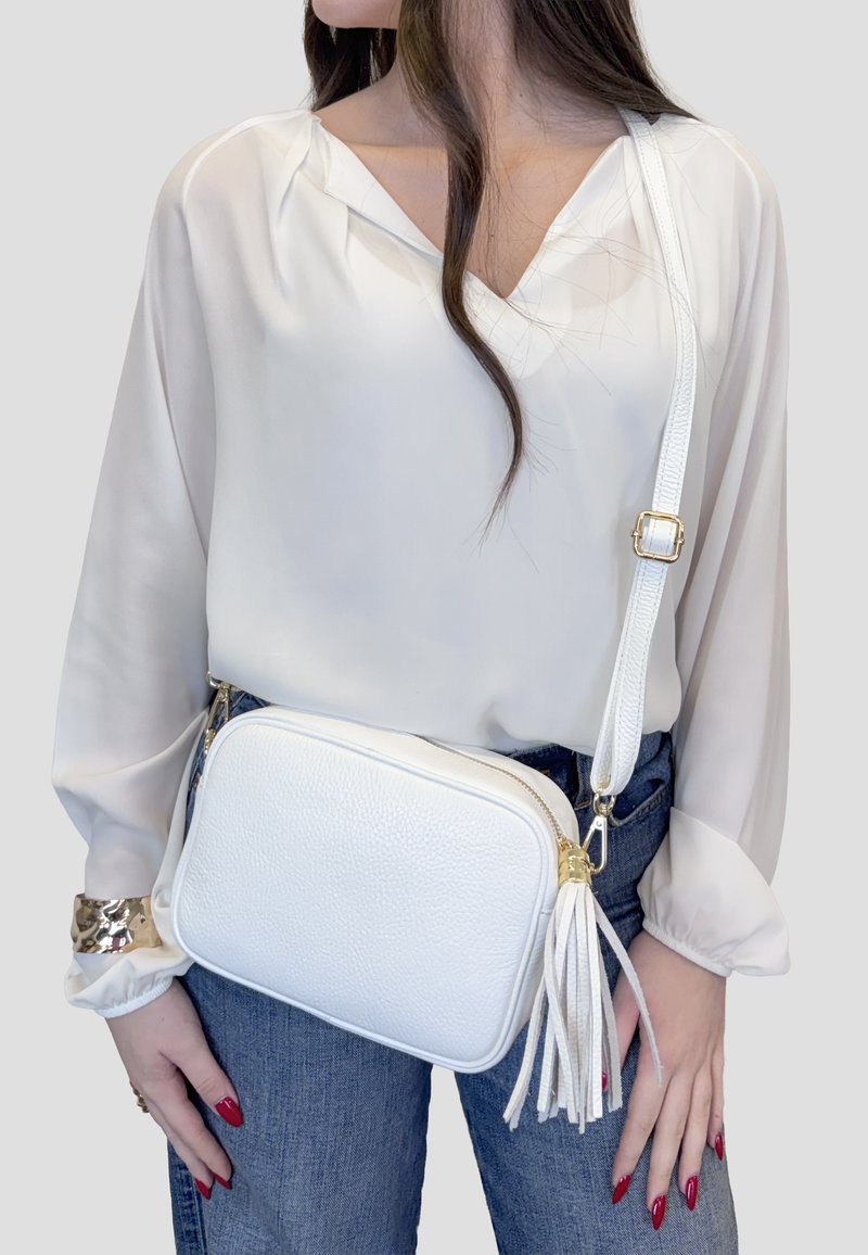 Vrouw draagt een wit blouse met lange mouwen en blauwe spijkerbroek, met een witte leren crossbodytas met kwastje en gouden gesp.