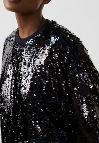 Persoon die een zwarte, glanzende top bedekt met pailletten draagt, met een ronde halslijn, waarbij de schouder en een deel van de nek zichtbaar zijn tegen een effen achtergrond.