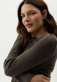 Brauner Strickpullover mit langen Ärmeln, der einen lockeren Schnitt und eine weiche Textur aufweist. Das Model posiert mit verschränkten Armen vor einem hellen Hintergrund.