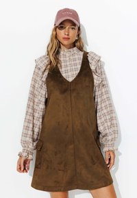 Robe salopette en daim brun sur un chemisier à carreaux crème et violet avec des manches volantées, associée à une casquette en velours côtelé rose.