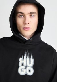 Sort hoodie med en frontkangaroo-lomme, der har et hvidt logo "HUGO" i flammedesign. Blødt stof, justerbar snor til hætten.