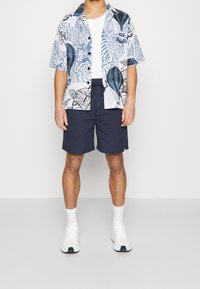 Camisa de botones de manga corta con estampado floral azul, combinada con pantalones cortos navy con cordón y zapatillas blancas, presentando un corte clásico.