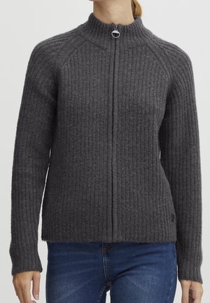 Woman wearing a dark gray ribbed zip-up sweater with long sleeves and high collar, paired with blue jeans, against a plain background.