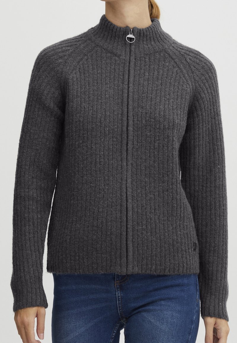 Donna che indossa un maglione a zip grigio scuro a coste con maniche lunghe e colletto alto, abbinato a jeans blu, su uno sfondo neutro.