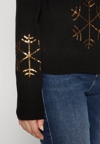 Pull noir avec motifs de flocons de neige en sequins dorés sur les manches, doté de poignets côtelés et d'une texture douce, associé à un jeans bleu.