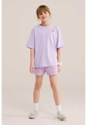 Nooruke helepruuni juustega, kandes lavendlivärvi üle-suuruses t-särki, triibulisi lühikeseid pükse, valgeid sokke ja halli spordijalatsit, seisab lõdvestunult.