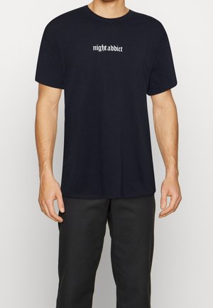 Mężczyzna w czarnej koszulce z białym gotyckim napisem "nightaddict" na środku klatki piersiowej, w połączeniu z czarnymi spodniami.