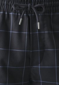 Bañador negro de tejido ligero con un patrón a cuadros en azul. Cuenta con una cintura elástica y cordón de ajuste con puntas de metal.