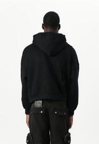 Sort cropped hoodie med snoretræk i hætten, lavet af blødt stof. Båret med mørke bukser, der har cargo-lommer og lynlåsdetaljer.