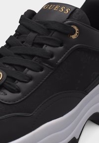 Scarpa da ginnastica nera Guess con logo dorato sulla linguetta, lacci neri, tomaia liscia e testurizzata, e suola imbottita bianca.