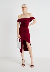 Robe bordeaux décolletée avec un design froncé et une fente sur le côté. Associée à un sac à main argenté et à des talons hauts à lanières. Texture de tissu lisse.