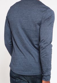 Muž oblečený v přiléhavé modré melírované košili s dlouhým rukávem a tmavých džínech, focený zezadu na jednobarevném bílém pozadí.