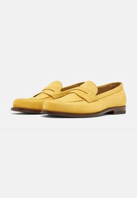 Mocasines de ante amarillos con detalle de tira cosida, suela de cuero marrón plana y diseño de punta redonda. Textura suave y estilo minimalista.