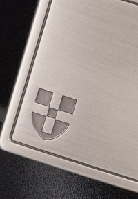 Prostokątny metalowy znacznik o szczotkowanym wykończeniu. Zawiera wygrawerowane logo z wzorem tarczy i elementami w kratkę. Ciemne tło.