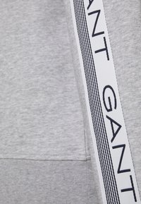 Felpa grigia con una tasca e un dettaglio bianco con strisce nere e la scritta "GANT". Tessuto morbido, design casual.