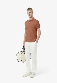 Brun polo med krage och grön logotyp, i kombination med vita byxor och en läderväska i beige med svarta accenter. Vita sneakers fulländar looken.