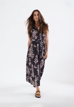 Mujer con cabello largo y castaño que lleva un vestido maxi negro sin mangas con un motivo de hojas en rosa claro y sandalias marrones, caminando hacia adelante sobre fondo blanco.