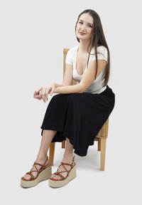 Bruine spletige sleehakken met een geweven jute basis, gedragen met een zwarte rok en een witte getailleerde blouse, zittend op een houten stoel.