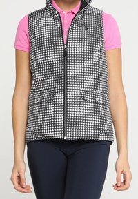 Svart och vit houndstooth-mönstrad väst med dragkedja framtill, hög krage och två sidofickor. Bärs över en rosa polo-tröja.