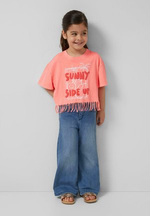 Uśmiechnięta dziewczyna o ciemnych włosach ma na sobie koralowy t-shirt z frędzlami z napisem „Sunny Side Up”, szerokie niebieskie dżinsy i sandały, stoi na tle szarej ściany.