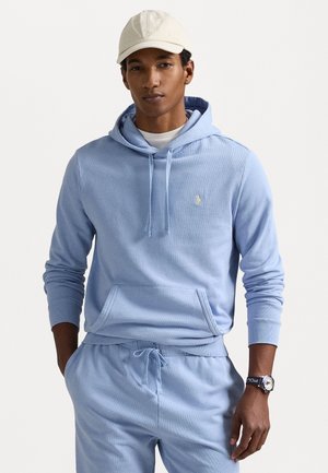 Hanorac de culoare albastru deschis, fabricat dintr-un material moale, având un buzunar tip cangur și un șnur. Este purtat cu pantaloni asortați și un tricou alb dedesubt.