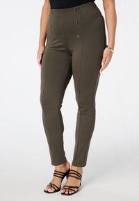 Femme portant un pantalon taille haute olive avec des boutons décoratifs et des sandales à talons noires à lanières, se tenant devant un fond blanc uni.