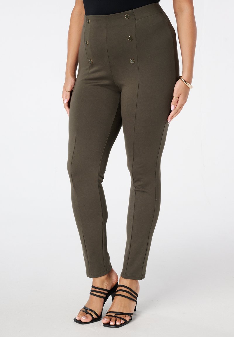 Femme portant un pantalon taille haute olive avec des boutons décoratifs et des sandales à talons noires à lanières, se tenant devant un fond blanc uni.