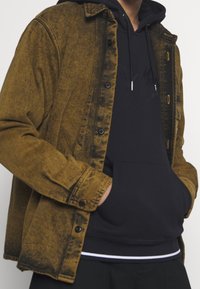 Mörkbrun denimjacka med en urblekt textur, knappad framsida och bröstfickor, lagd över en svart hoodie med en kenguruficka.