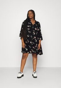 Black floral dress with a V-neck, tiered design, and ruffled sleeves, featuring a pink flower pattern. Paired with white shoes.