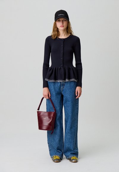 Donna con cardigan peplum nero abbottonato, jeans blu a gamba larga, berretto nero, sneakers con lacci gialli, che tiene una borsa bordeaux al fianco.