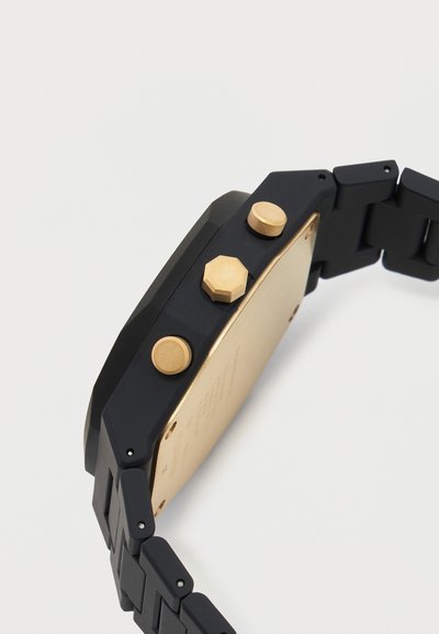 Μαύρο έξυπνο ρολόι με χρυσή πίσω πλάκα και τρία χρυσά πλευρικά κουμπιά, συνδεδεμένο με μαύρο ρολόι με κρίκους, εμφανιζόμενο σε γωνία σε λευκό φόντο.