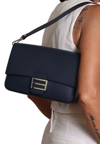 Vrouw in een witte mouwloze top en broek, die een marineblauwe rechthoekige handtas met een zilveren gesp op haar schouder draagt.