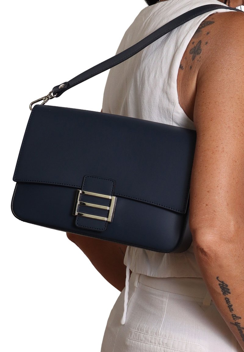 Vrouw in een witte mouwloze top en broek, die een marineblauwe rechthoekige handtas met een zilveren gesp op haar schouder draagt.