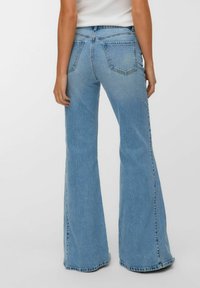 Vrouw in lichtblauwe high-waisted flare jeans en een witte mouwloze top, gezien van achteren tegen een effen achtergrond.