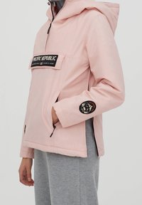 Lyselilla anorak-jakke med en front kangaroo lomme, sort mærke med teksten "PACIFIC REPUBLIC" og et cirkulært badge på ærmet.