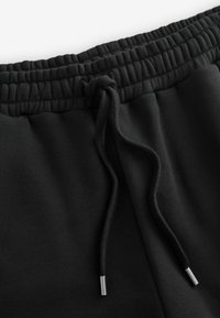 Zwarte joggingbroek met een gefronst elastieken tailleband en trekkoord, gemaakt van soepel materiaal met contrasterende metalen punten op het trekkoord.