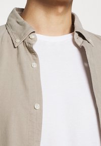 Camisa de pana color beige claro con cuello abotonado, con botones blancos y una camiseta blanca lisa debajo.