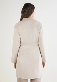 Beżowy płaszcz o średniej długości z kołnierzem, długimi rękawami i wiązanym w pasie. Gładka tekstura z prostym, minimalistycznym wzornictwem.