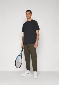 Hall halli lühikeste varrukatega T-särk, oliivrohelised dressipüksid ja valged spordijalatsid; hoiab käes musta-valget tennise reketit.