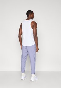 Άνδρας με λευκό αμάνικο μπλουζάκι, ανοιχτό μωβ παντελόνι jogger, λευκές κάλτσες και λευκά αθλητικά παπούτσια, στέκεται μπροστά σε έναν απλό ανοιχτό γκρι τοίχο.